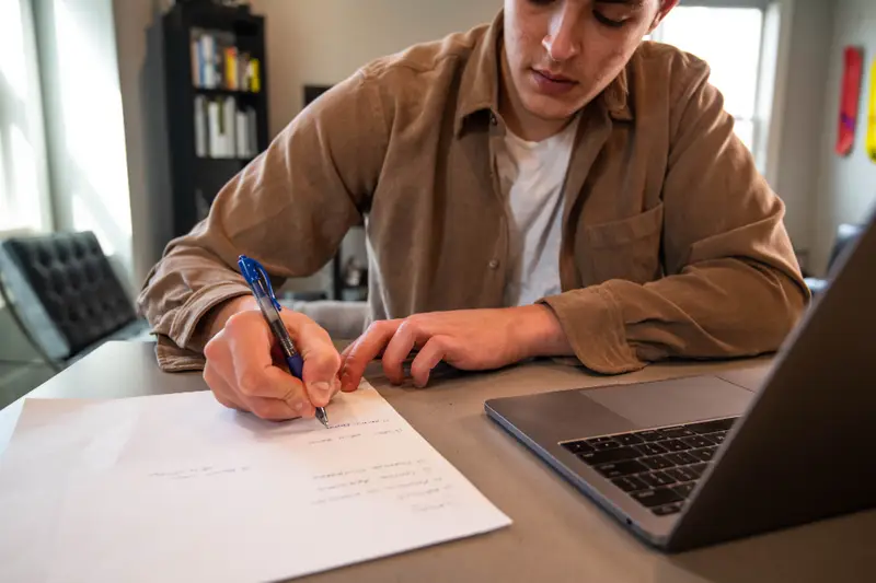 Hombre escribiendo en un papel junto a un portátil