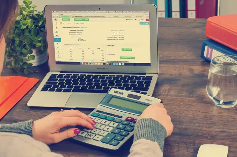 Persona usando una calculadora junto a una hoja de cálculo en un portátil